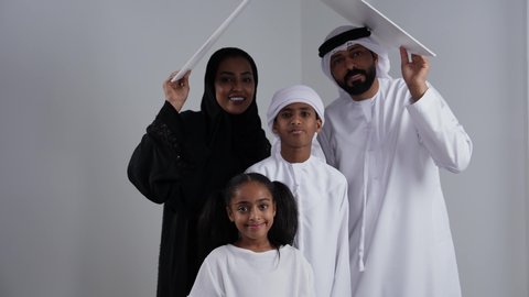 تلبية الاحتياجات المنزليه العائلية، ايماءات تدل على السعادة, الاستقرار العائلي في الاسكان والتمليك ، بورتريه لزوجان عربيان خليجيان اماراتيان يرتديان الزي التقليدي يحملان لوح أبيض فوق رأس الابناء, قضاء الوقت مع الأهل والأبناء ، خلفية بيضاء
