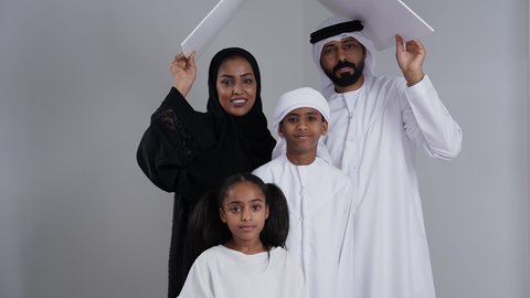 قضاء الوقت مع الأهل والأبناء, الاستقرار العائلي في الاسكان والتمليك, ايماءات تدل على السعادة, تلبية الاحتياجات المنزليه العائلية، بورتريه لزوجان عربيان خليجيان اماراتيان يرتديان الزي التقليدي يحملان لوح أبيض فوق رأس الابناء، خلفية بيضاء