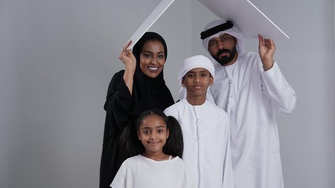 Family stability in housing and ownership, gestures indicating happiness, meeting family home needs, a portrait of an Emirati Gulf Arab couple wearing traditional attire holding a white board above their children's heads, spending time with family and children, white background.