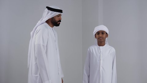 الاهتمام بالاولاد وتقديم الدعم والرعاية، مفهوم الرابطة الأبوية والترابط الأسري، ايماءات الحب والعطف على الأبناء، بورتريه لرجل عربي خليجي اماراتي يرتدي الكندورة والغترة يسمك بيد ابنه، النظر للكاميرا بتعابير السعادة والسرور، خلفية بيضاء