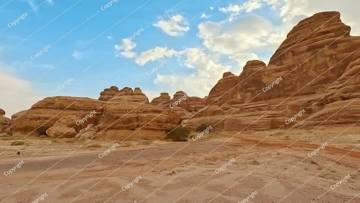 تشكيلات صحراوية صخرية مع رمال في المقدمة، سماء زرقاء مع سحب في الأعلى.