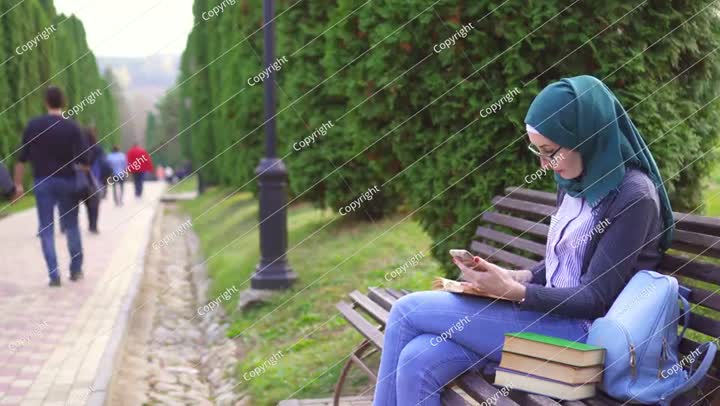طالب مسلم يحمل هاتفًا وكتابًا على مقعد في الحديقة عند غروب الشمس