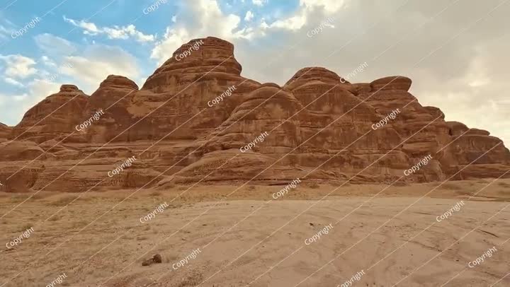Rocky desert formations with sand in foreground, blue sky with clouds above..
