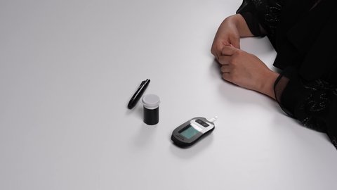 استخدام الأجهزة الالكترونية للتحاليل المنزلية، الفحوصات الطبية السريعة لمعرفة نسبة السكر في الدم، شابة سعودية مصابة بحالة مرضية مزمنة, المتابعة الدورية لمرضى داء السكري, تصوير مقرب لامرأة عربية خليجية إماراتية ترتدي العباءة تقوم بوخز اصبعها, خلفية بيضاء