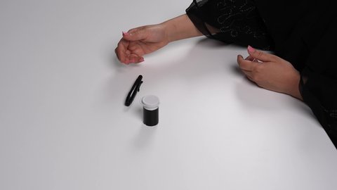 الفحوصات الطبية السريعة لمعرفة نسبة السكر في الدم، استخدام الأجهزة الالكترونية للتحاليل المنزلية، شابة سعودية مصابة بحالة مرضية مزمنة, المتابعة الدورية لمرضى داء السكري, تصوير مقرب لامرأة عربية خليجية إماراتية ترتدي العباءة تقوم بوخز اصبعها, خلفية بيضاء