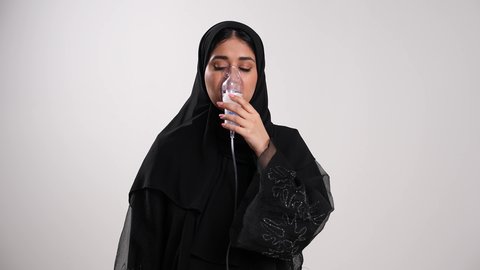 الشعور بالاختناق وضيق في النفس، استعمال قناع التنفس الصناعي، إمرأة سعودية مصابة بتضرر في مجرى الهواء، أجهزة استنشاق خاصة بمرضى الجهاز التنفسي والرئتين، تصوير مقرب لشابة عربية خليجية إماراتية محجبة ترتدي العباءة تعاني من أزمة تنفس تحسسية، خلفية بيضاء