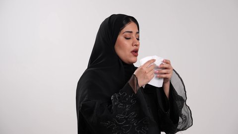 الشعور بالتعب والإرهاق والمرض، تصوير مقرب من الجانب لامرأة عربية خليجية إماراتية محجبة ترتدي العباءة السوداء تقوم بالعطاس، شابة سعودية مصابة بالزكام والرشح، الإصابة بنزلات البرد، ظهور أعراض فايروس كورونا، استعمال المناديل الورقية أثناء العطس،خلفية بيضاء