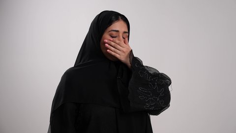 تصوير مقرب لشابة عربية خليجية إماراتية محجبة ترتدي العباءة تقوم بالتثاؤب، إيماءات التعب والإرهاق والحاجة لأخذ قسط من الراحة، أهمية الخلود للنوم باكرا واتباع نظام صحي، الإحساس بالملل والخمول، امرأة سعودية تضع يدها أمام فمها بتعابير النعاس والكسل، خلفية بيضاء