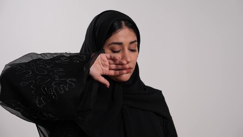 A Saudi woman feels sick and exhausted, gestures of fatigue and the need for rest, the importance of visiting a doctor and taking the appropriate treatment, using an oral thermometer to determine body temperature, a close-up shot of a young Emirati Gulf Arab woman wearing a hijab and an abaya measuring her temperature with a digital thermometer, white background.