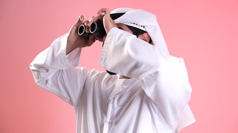 تصوير مقرب لشاب عربي خليجي اماراتي يرتدي الكندورة والغترة ينظر من خلال المنظار ثنائي العينين بايماءات الاستغراب، حب المغامرة والاستكشاف، البحث عن شيء ما وتوسيع مجال الرؤية، رؤية الاشياء البعيدة عبر التلسكوب، التخطيط المستقبلي للأهداف والطموحات، خلفية وردية