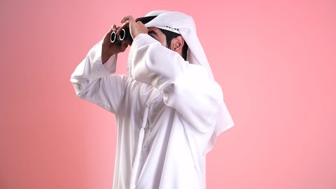 التخطيط المستقبلي للأهداف والطموحات، تصوير مقرب لشاب عربي خليجي اماراتي يرتدي الكندورة والغترة ينظر من خلال المنظار ثنائي العينين بايماءات الاستغراب، حب المغامرة والاستكشاف، البحث عن شيء ما وتوسيع مجال الرؤية، رؤية الاشياء البعيدة عبر التلسكوب، خلفية وردية