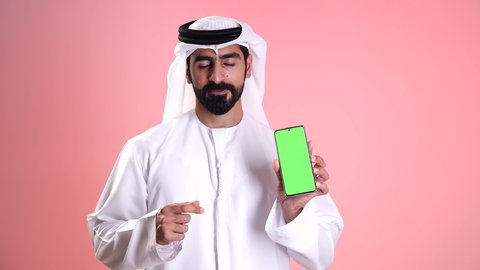 عرض شاشة خضراء فارغة عبر الجوال, النظر إلى الكاميرا بإيماءات الإعجاب والرضى, استخدام الأجهزة التقنية الحديثة في الحياة اليومية, تصوير مقرب لشاب عربي خليجي إماراتي يرتدي الكندورة والغترة يمسك بيده الهاتف المحمول ويشير بإصبع السبابة إلى الموبايل, خلفية وردية