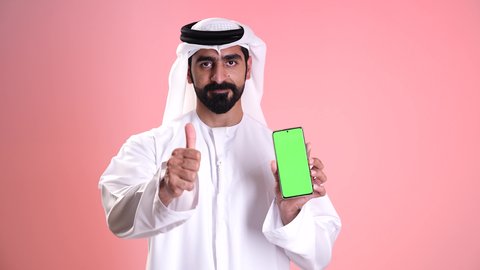 النظر إلى الكاميرا بإيماءات الإعجاب والرضى, عرض شاشة خضراء فارغة عبر الجوال,استخدام الأجهزة التقنية الحديثة في الحياة اليومية, تصوير مقرب لشاب عربي خليجي إماراتي يرتدي الكندورة والغترة يمسك بيده الهاتف المحمول ويشير بإصبع السبابة إلى الموبايل, خلفية وردية