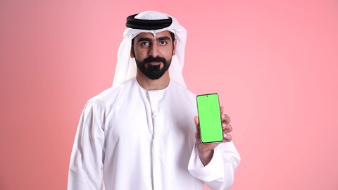 استخدام جهاز تقني حديث ومتطور، عرض شاشة خضراء فارغة عبر الجوال, النظر إلى الكاميرا بإيماءات الإعجاب والرضى, تصوير مقرب لشاب عربي خليجي إماراتي يرتدي الكندورة والغترة يمسك بيده الهاتف المحمول ويشير بإصبع السبابة إلى الموبايل, خلفية وردية
