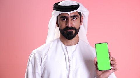 تصوير مقرب لشاب عربي خليجي إماراتي يرتدي الكندورة والغترة يمسك بيده الهاتف المحمول ويشير بإصبع السبابة إلى الموبايل, عرض شاشة خضراء فارغة عبر الجوال, النظر إلى الكاميرا بإيماءات الإعجاب والرضى, استخدام الأجهزة التقنية الحديثة في الحياة اليومية, خلفية وردية