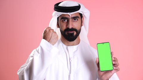 عرض شاشة خضراء فارغة عبر الجوال, النظر إلى الكاميرا بإيماءات الإعجاب والرضى, استخدام الأجهزة التقنية الحديثة في الحياة اليومية, تصوير مقرب لشاب عربي خليجي إماراتي يرتدي الكندورة والغترة يمسك بيده الهاتف المحمول ويشير بإصبع السبابة إلى الموبايل, خلفية وردية