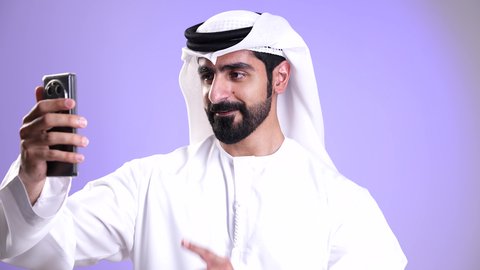 إجراء مكالمة فيديو مع الأهل والأصدقاء عبر الهاتف المحمول، ايماءات السلام والترحيب, تصوير مقرب لشاب عربي خليجي إماراتي يرتدي الكندورة والغترة يقوم ببث مباشر على مواقع التواصل، استخدام التقنيات الحديثة والمتطورة, تبادل الأخبار والأحاديث, خلفية موف