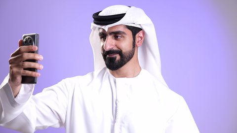 استخدام التقنيات الحديثة والمتطورة, إجراء مكالمة فيديو مع الأهل والأصدقاء عبر الهاتف المحمول، ايماءات السلام والترحيب, تصوير مقرب لشاب عربي خليجي إماراتي يرتدي الكندورة والغترة يقوم ببث مباشر على مواقع التواصل، تبادل الأخبار والأحاديث, خلفية موف