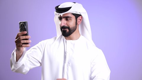 إجراء مكالمة فيديو مع الأهل والأصدقاء عبر الهاتف المحمول، ايماءات السلام والترحيب, تصوير مقرب لشاب عربي خليجي إماراتي يرتدي الكندورة والغترة يقوم ببث مباشر على مواقع التواصل، استخدام التقنيات الحديثة والمتطورة, تبادل الأخبار والأحاديث, خلفية موف