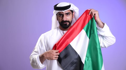 يوم العلم الاماراتي 3 نوفمبر، الفخر بالانتماء للوطن، الاعياد والمناسبات الوطنية، ذكرى استقلال الإمارات العربية المتحدة في 2 ديسمبر، تصوير مقرب لرجل عربي خليجي اماراتي يرتدي الكندورة والغترة يحمل علم الإمارات ويرفع اصبع الابهام بعلامة الامتياز، خلفية موف