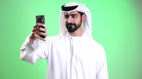 Communication with family and friends, gestures of peace and welcome, a close-up shot of a young Emirati Arab man wearing a kandura and ghutrah making a video call via mobile phone, using modern and advanced technologies, exchanging news and conversations, live broadcasting on social media, green background, chroma.
