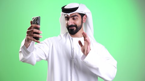 Gestures of peace and welcome, communication with family and friends, a close-up shot of a young Emirati Arab man wearing a kandura and ghutrah making a video call via mobile phone, using modern and advanced technologies, exchanging news and conversations, live broadcasting on social media, green background, chroma.