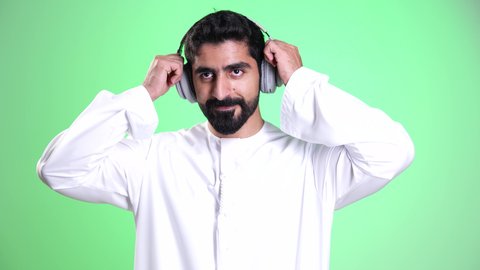 استخدام تقنية الهيدفون، مفهوم الإصغاء والاستماع, قضاء الأوقات الممتعة بسماع الأغاني المفضلة, استخدام الأجهزة التقنية الحديثة للتسلية والترفيه, تصوير مقرب لشاب عربي خليجي إماراتي يرتدي الكندورة والغترة يضع سماعات الرأس الصوتية, خلفية خضراء، كروما