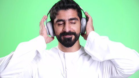 مفهوم الإصغاء والاستماع, استخدام تقنية الهيدفون، قضاء الأوقات الممتعة بسماع الأغاني المفضلة, استخدام الأجهزة التقنية الحديثة للتسلية والترفيه, تصوير مقرب لشاب عربي خليجي إماراتي يرتدي الكندورة والغترة يضع سماعات الرأس الصوتية, خلفية خضراء، كروما