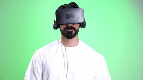 Interacting with the imaginary world, enjoying watching something with 3D VR glasses, a close-up shot of an Emirati Gulf Arab man wearing a kandura and ghutrah simulating virtual reality, gestures of immersion and amazement, the concept of technological evolution, chroma, green background.