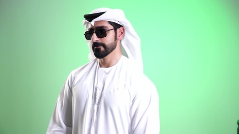 White Friday discounts, a close-up shot of a smiling Emirati Gulf Arab young man wearing a kandura and ghutrah, wearing sunglasses and holding paper shopping bags in his hand, looking at the camera with expressions of joy and excitement, acquiring supplies and gifts, green background, chroma.