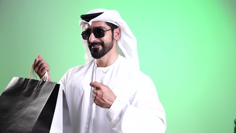 Looking at the camera with expressions of joy and excitement, taking advantage of seasonal offers and discounts, a close-up shot of a smiling young Emirati Arab man wearing a kandura and ghutrah, wearing sunglasses and holding paper shopping bags in his hand, acquiring supplies and gifts, green background, chroma.