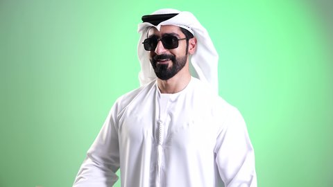 اقتناء المستلزمات والهدايا، الاستفادة من العروض الموسمية والتخفيضات، تصوير مقرب لشاب عربي خليجي إماراتي مبتسم يرتدي الكندورة والغترة ويضع النظارة الشمسية يحمل بيده أكياس التسوق الورقية، النظر الى الكاميرا بتعابير السرور والحماس، خلفية خضراء، كروما