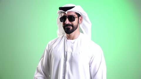 الاستفادة من العروض الموسمية والتخفيضات، تصوير مقرب لشاب عربي خليجي إماراتي مبتسم يرتدي الكندورة والغترة ويضع النظارة الشمسية يحمل بيده أكياس التسوق الورقية، النظر الى الكاميرا بتعابير السرور والحماس، اقتناء المستلزمات والهدايا، خلفية خضراء، كروما