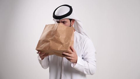 تأمين الأوكسجين بشكل عاجل للرئتين، الأمراض والمشاكل التحسسية الصدرية، اتباع إجراءات استنشاق خاصة بمرضى الجهاز التنفسي، الشعور بالهلع وضيق في التنفس، تصوير مقرب لشاب عربي خليجي إماراتي يرتدي الكندورة والغترة يتنفس في كيس ورقي، خلفية بيضاء