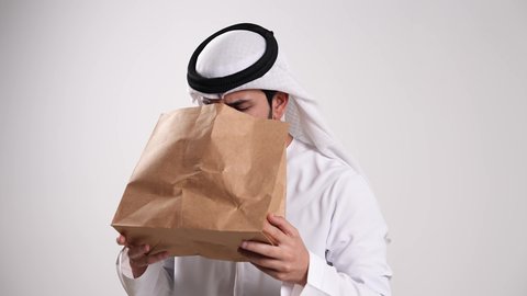 الأمراض والمشاكل التحسسية الصدرية، تأمين الأوكسجين بشكل عاجل للرئتين، اتباع إجراءات استنشاق خاصة بمرضى الجهاز التنفسي، الشعور بالهلع وضيق في التنفس، تصوير مقرب لشاب عربي خليجي إماراتي يرتدي الكندورة والغترة يتنفس في كيس ورقي، خلفية بيضاء