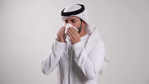 المعاناة من التهابات الأنف التحسسية، الإصابة بالانفلونزا ونزلات البرد، ظهور أعراض كوفيد_ ١٩ ، حالات الزكام والرشح، الشعور بالتعب والإرهاق والحاجة للراحة، تصوير مقرب لشاب عربي خليجي إماراتي يرتدي الكندورة والغترة يمسك بمنديل ورقي ويقوم بالعطاس، خلفية بيضاء