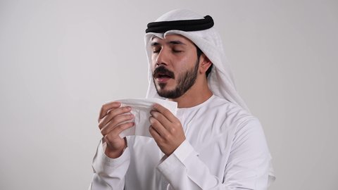الإصابة بالانفلونزا ونزلات البرد، المعاناة من التهابات الأنف التحسسية، ظهور أعراض كوفيد_ ١٩ ، حالات الزكام والرشح، الشعور بالتعب والإرهاق والحاجة للراحة، تصوير مقرب لشاب عربي خليجي إماراتي يرتدي الكندورة والغترة يمسك بمنديل ورقي ويقوم بالعطاس، خلفية بيضاء