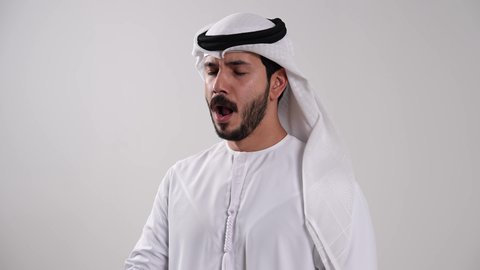 أهمية الخلود للنوم باكرا واتباع نظام صحي، وضع اليد أمام الفم بتعابير النعاس والكسل،إيماءات التعب والإرهاق والحاجة لأخذ قسط من الراحة، الإحساس بالملل والخمول، تصوير مقرب لشاب عربي خليجي إماراتي يرتدي الكندورة والغترة يقوم بالتثاؤب، خلفية بيضاء