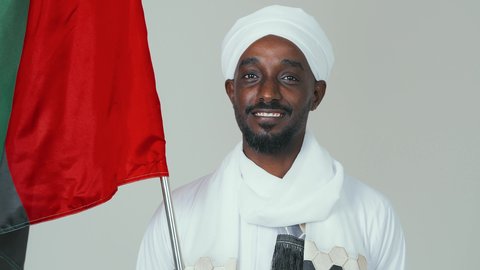 حمل العلم السوداني بفخر،الاحتفال بالاستقلال ١ يناير، استذكار اليوم الوطني السوداني ١٩٥٦ م، ايماءات الاعتزاز والثقة،  حب الوطن والاعتزاز به، بورتريه مقرب لرجل عربي سوداني يرتدي التوب والشال والعمه البيضاء يقف بثقة واستقامة، خلفية بيضاء