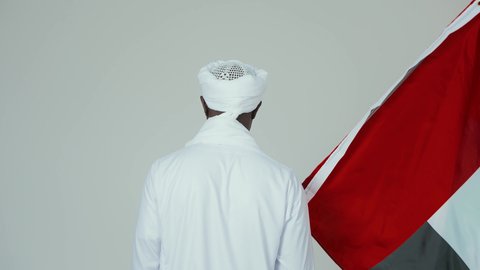 مفهوم الانتماء الوطني، رفرفه العلم السوداني بفخر،الاحتفال بالاستقلال ١ يناير، استذكار اليوم الوطني السوداني ١٩٥٦ م، ايماءات الاعتزاز والثقة،  حب الوطن والاعتزاز به، بورتريه خلفي لرجل عربي سوداني يرتدي التوب والشال والعمه البيضاء يقف بثقة واستقامة، خلفية بيضاء