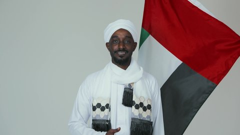 رفع اصبع الابهام بإيماءات الاعجاب، حمل العلم السوداني بفخر،الاحتفال بالاستقلال ١ يناير، استذكار اليوم الوطني السوداني ١٩٥٦ م، حب الوطن والاعتزاز به، بورتريه مقرب لرجل عربي سوداني يرتدي الجلابية والشال والعمتمه البيضاء يقف بثقة واستقامة، خلفية بيضاء