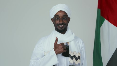Raising the thumb with gestures of admiration, proudly holding the Sudanese flag, celebrating independence on January 1, commemorating Sudan's National Day in 1956, love for the homeland and pride in it, a close portrait of a Sudanese Arab man wearing a jalabiya, shawl, and white turban standing confidently and upright, with a white background.
