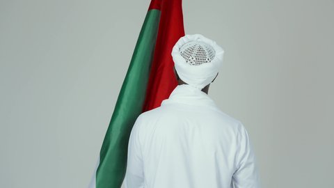 Love of the homeland and pride in it, celebrating independence on January 1, commemorating Sudan's National Day in 1956, a portrait from behind of a Sudanese Arab man wearing a jalabiya, shawl, and white turban, draping the Sudanese flag over his shoulders, with a white background.