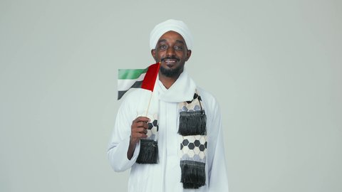 ايماءات السعادة والسرور، مفهوم الحب و الانتماء للبلاد, الأعياد و المناسبات الوطنية,  الاحتفال باليوم الوطني الإماراتي 2 ديسمبر, بورتريه لرجل عربي سوداني يرتدي العمامة والجلابية والشال يحمل علم الإمارات العربية المتحدة, خلفية بيضاء