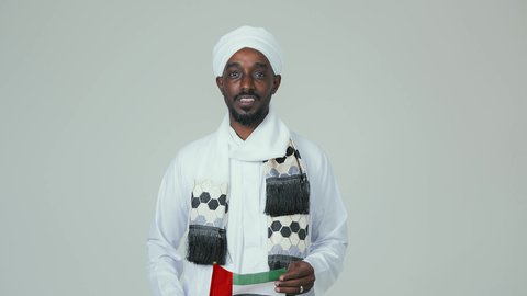 مفهوم الحب و الانتماء للبلاد, الاحتفال باليوم الوطني الإماراتي 2 ديسمبر ، ايماءات السعادة والسرور،  الأعياد و المناسبات الوطنية, بورتريه لرجل عربي سوداني يرتدي العمامة والجلابية والشال يحمل علم الإمارات العربية المتحدة, خلفية بيضاء