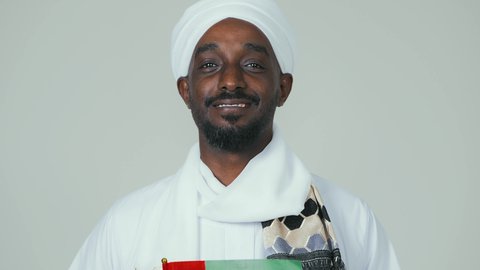 مفهوم الحب و الانتماء للبلاد, الاحتفال باليوم الوطني الإماراتي 2 ديسمبر ، ايماءات السعادة والسرور،  الأعياد و المناسبات الوطنية, بورتريه لرجل عربي سوداني يرتدي العمامة والجلابية والشال يحمل علم الإمارات العربية المتحدة, خلفية بيضاء