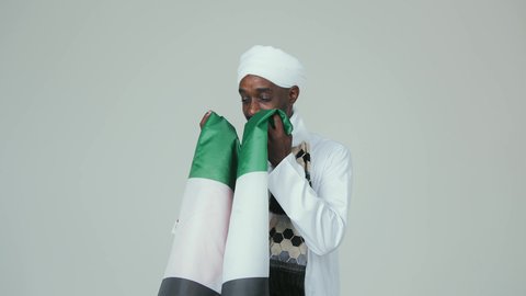 مفهوم الحب و الانتماء للبلاد, الاحتفال باليوم الوطني الإماراتي 2 ديسمبر ، ايماءات السعادة والسرور،  الأعياد و المناسبات الوطنية, بورتريه لرجل عربي سوداني يرتدي العمامة والجلابية والشال يقبل علم الإمارات العربية المتحدة, خلفية بيضاء