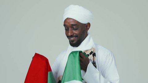 The concept of love and belonging to the country, celebrating the UAE National Day on December 2, gestures of happiness and joy, national holidays and occasions, a close-up portrait of a Sudanese Arab man wearing a turban, jalabiya, and shawl kissing the flag of the United Arab Emirates, white background.