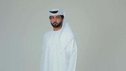 الوقوف بايدي متقاطعة ، الاناقة و الاهتمام  بالمظهر الخارجي، بورتريه لرجل عربي خليجي اماراتي يرتدي الكندورة و الغترة ينظر الى الكاميرا بايماءات الثقة بالنفس ، خلفية بيضاء