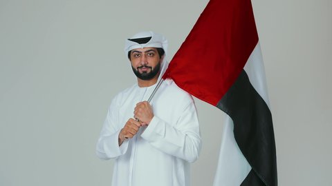 السعادة والسرور في المناسبات الوطنية، الاحتفال باليوم الوطني الإماراتي, الأعياد و المناسبات الوطنية,  مفهوم الحب و الانتماء للبلاد, بورتريه مقرب لرجل عربي خليجي إماراتي يرتدي الكندورة والغترة البيضاء يقوم بحمل علم الإمارات العربية المتحدة, خلفية بيضاء